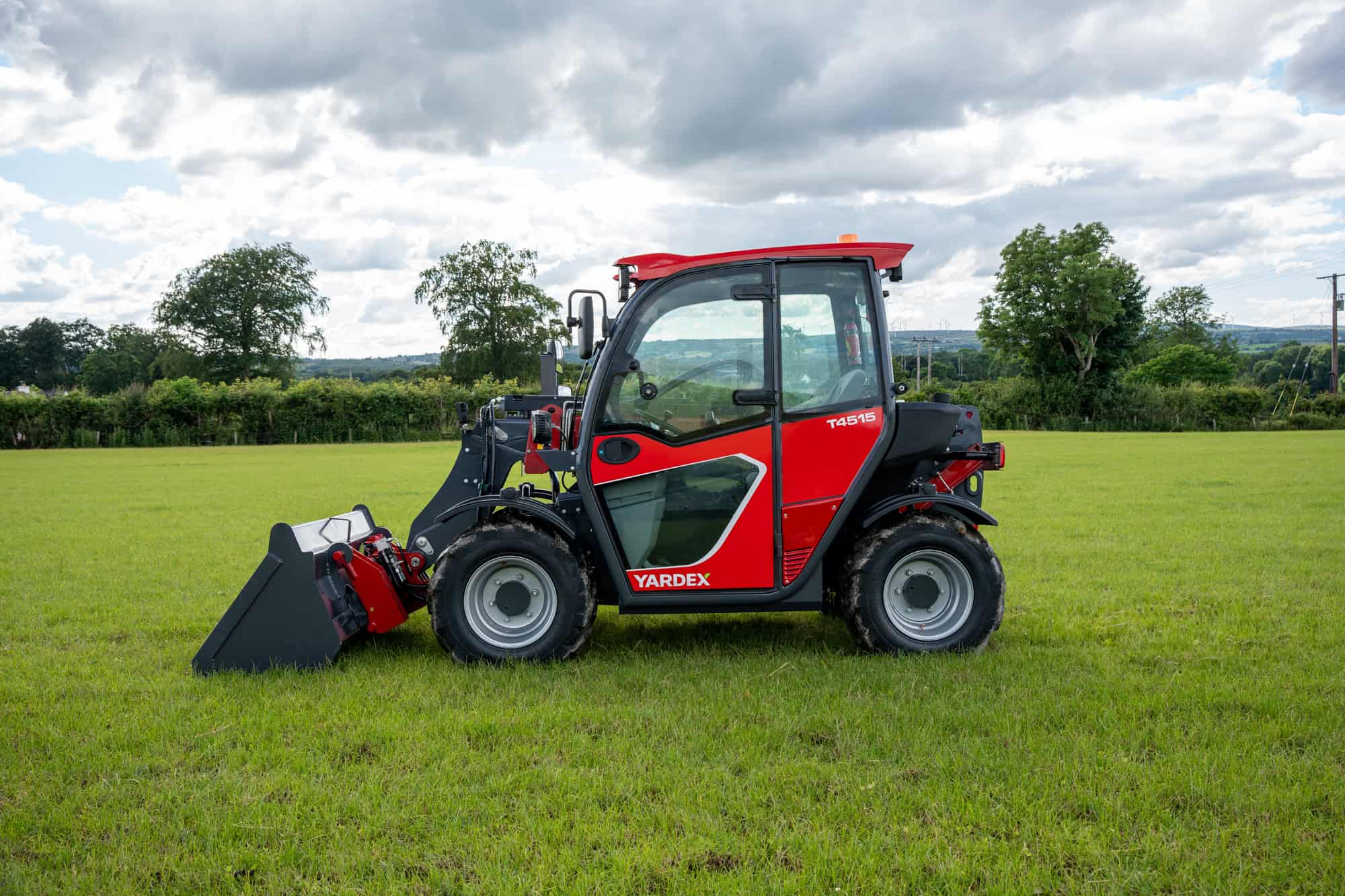 Yardex Group compact utility loader on green field with trees and cloudy sky.