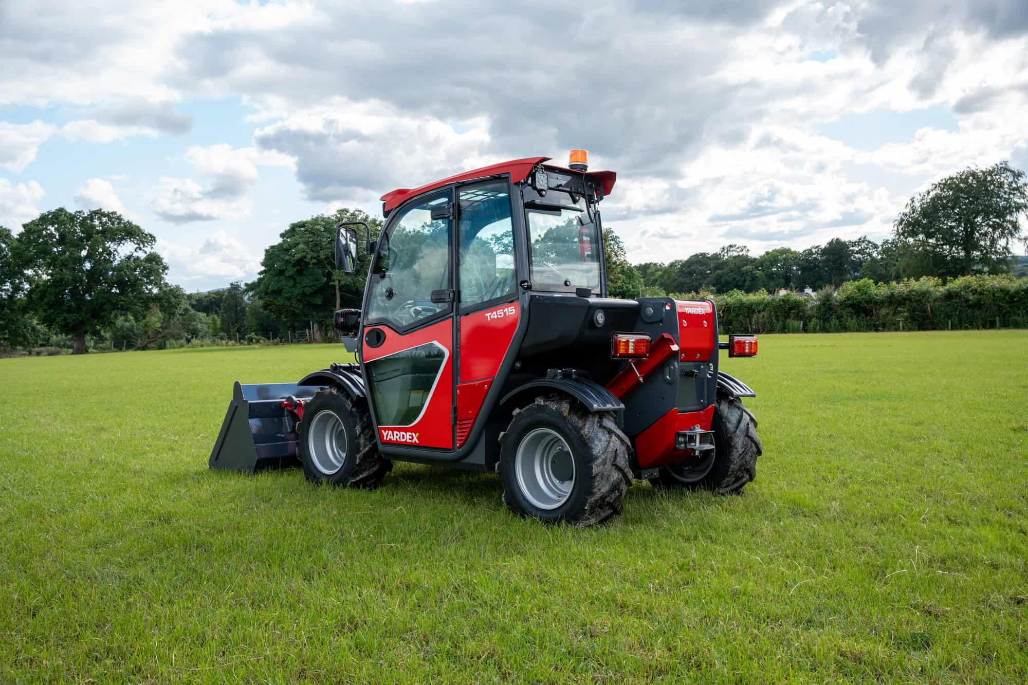 Red compact utility loader on green grass with trees and cloudy sky.