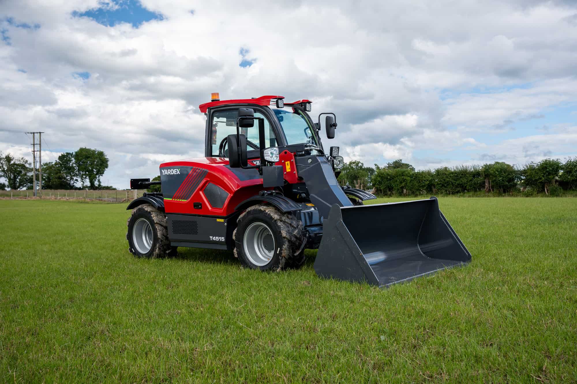 Yardex Group compact loader with bucket on grassy field under cloudy sky.