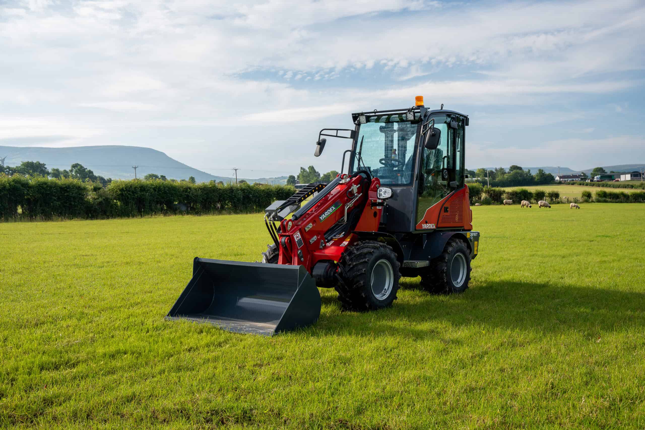 Yardex Group compact loader working on green field with mountains in background.