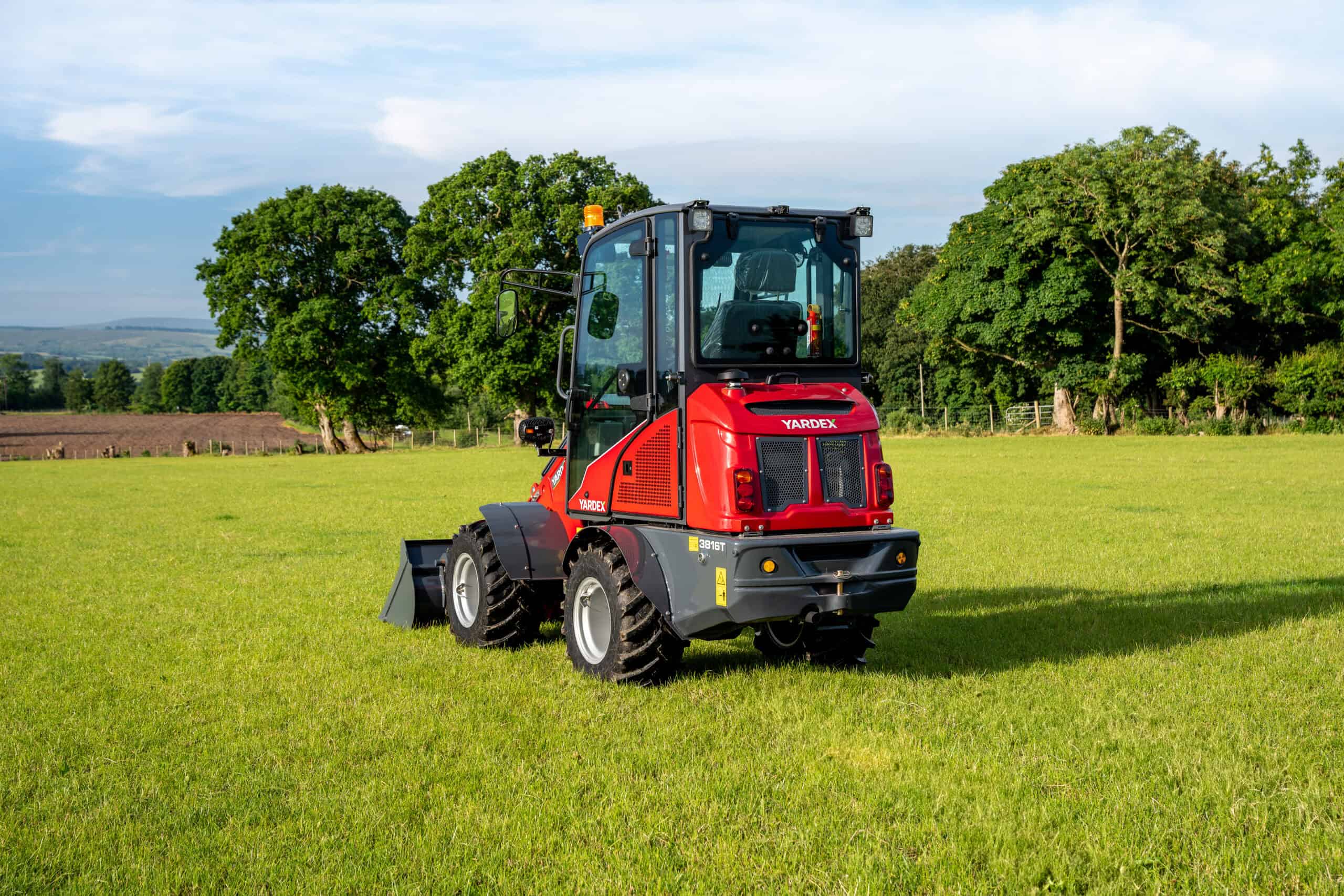 Red compact utility loader on green field with trees in background.