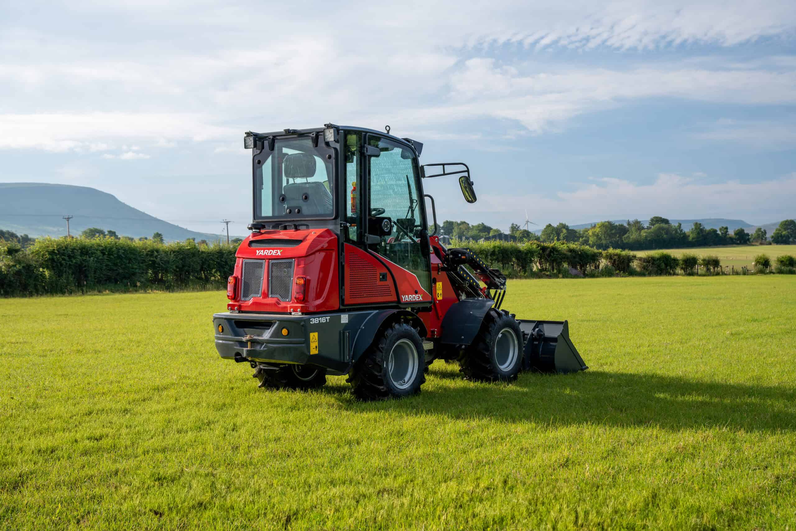 Red compact utility loader on green field with blue sky and mountains.