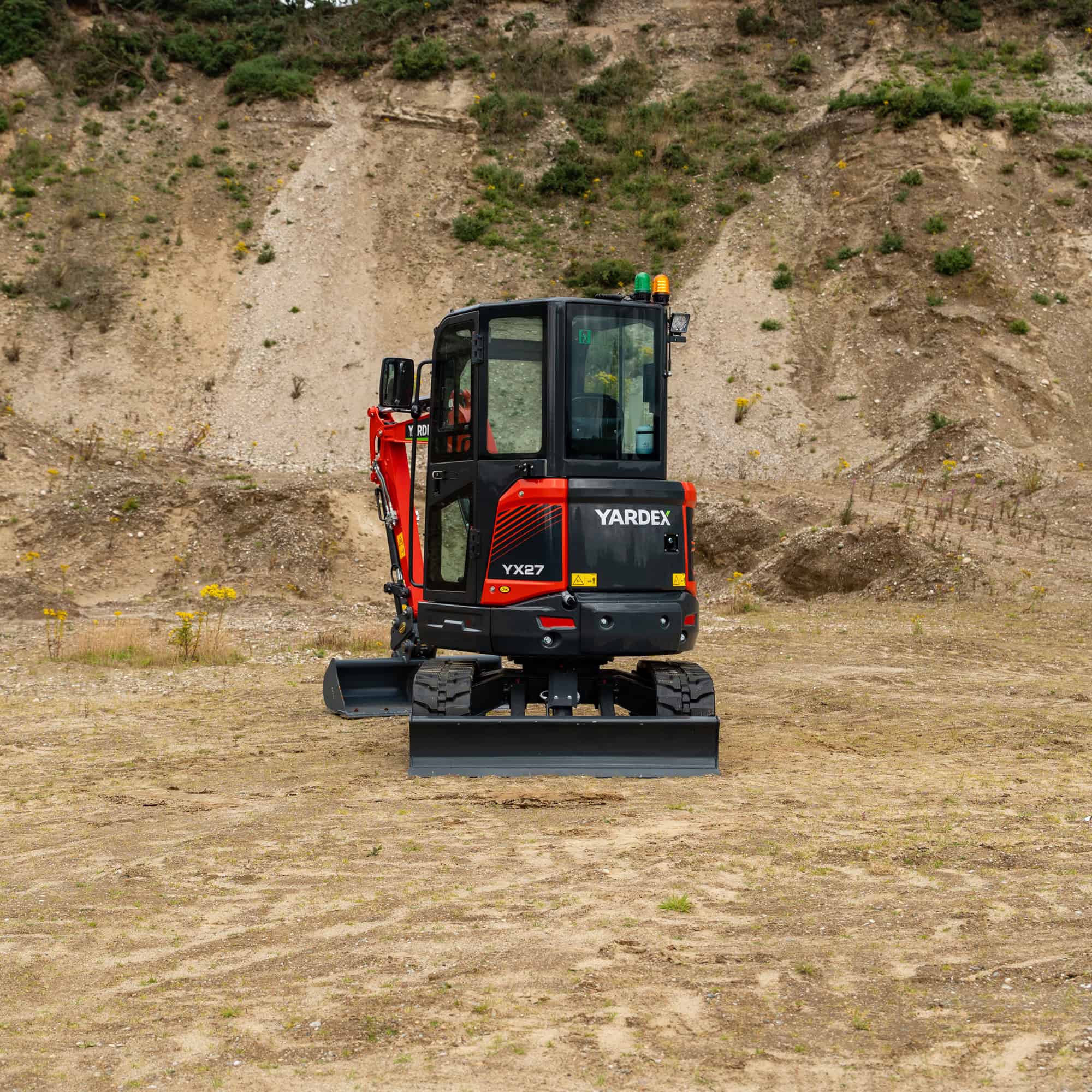 Yardex compact excavator working on a construction site with dirt and rocky terrain.