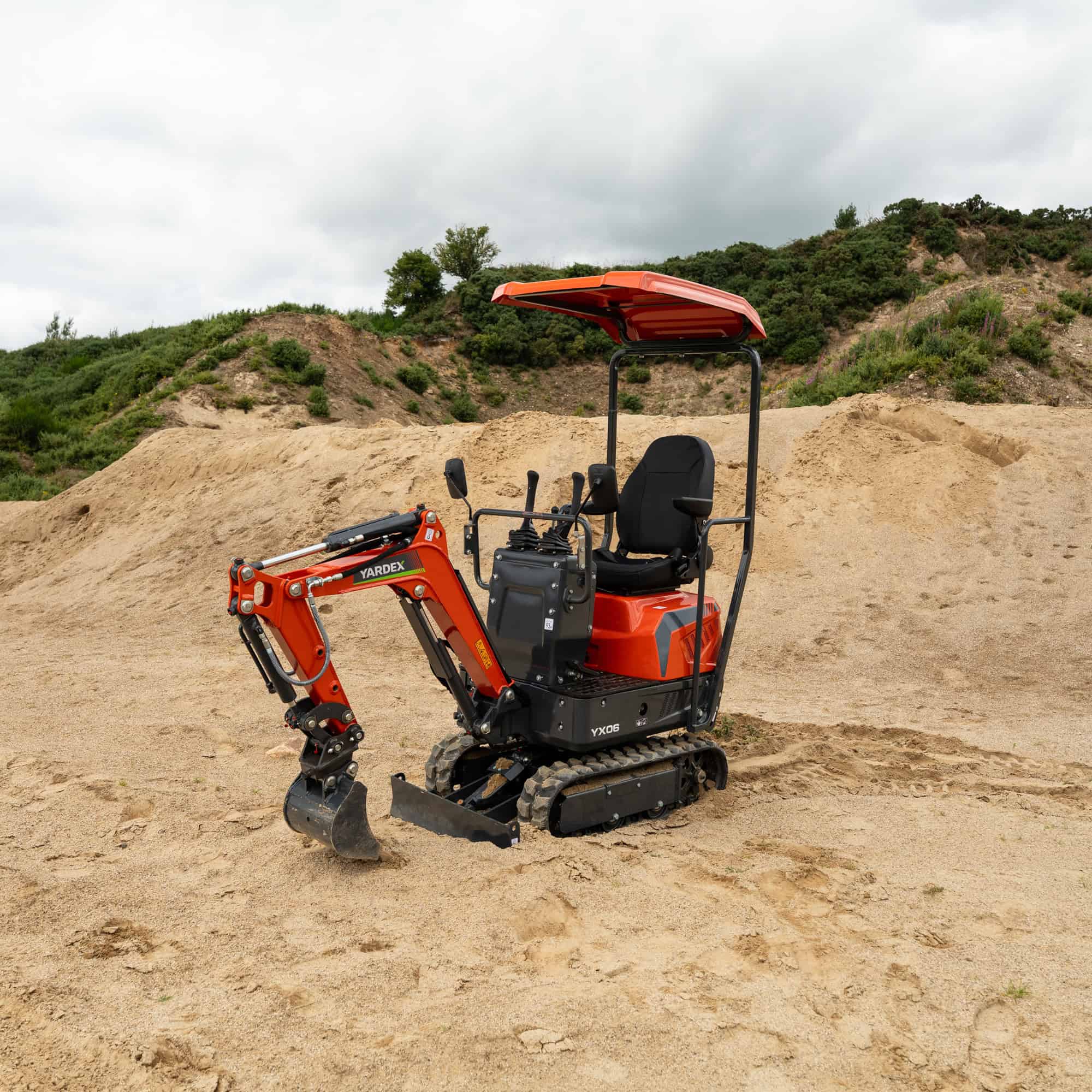 Compact orange Yardex mini excavator on sandy terrain with hills in background.