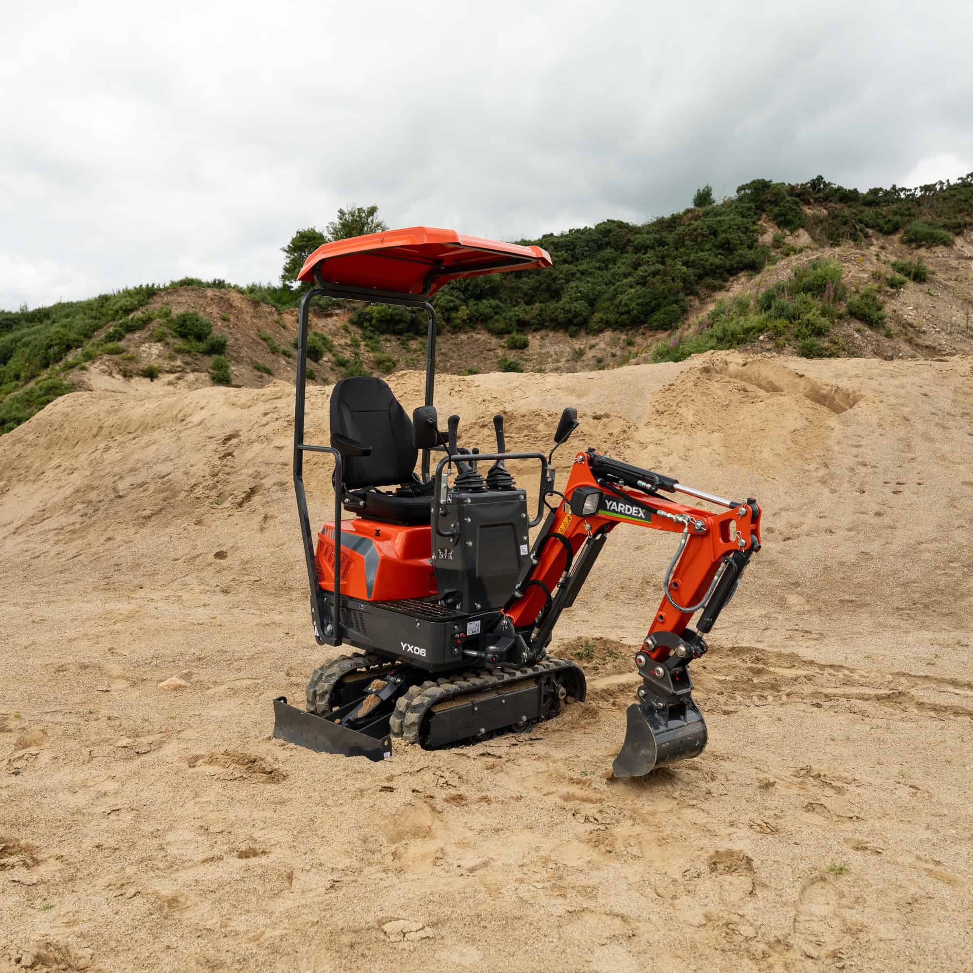 Compact orange Yardex mini excavator on sandy terrain with hills in background.